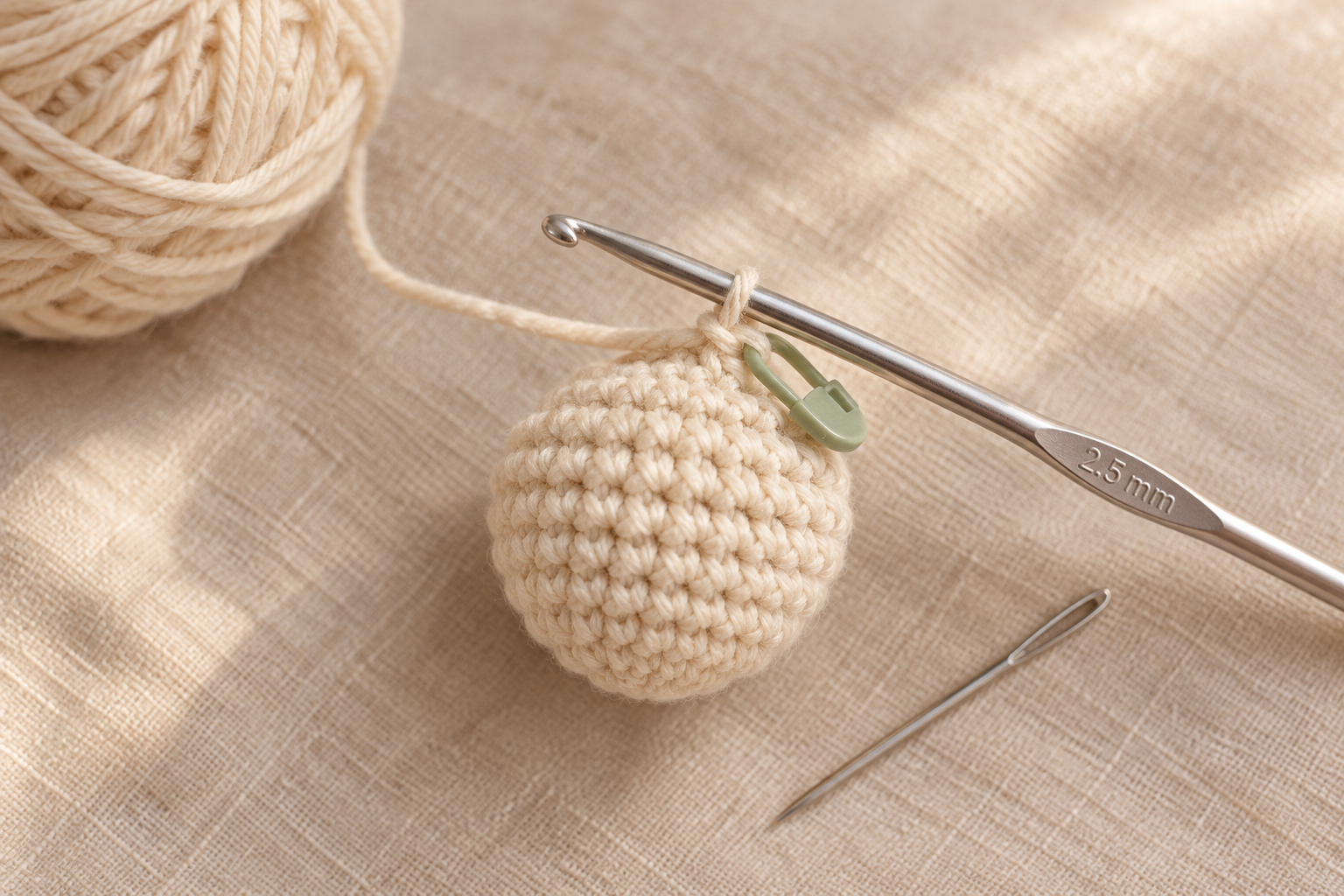 Overhead close-up of a small cream cotton crochet acorn body in progress at round 6 — a round cream ball on a 2.5 mm silver hook, small ball of cream cotton DK yarn softly blurred left, locking stitch marker clipped in place
