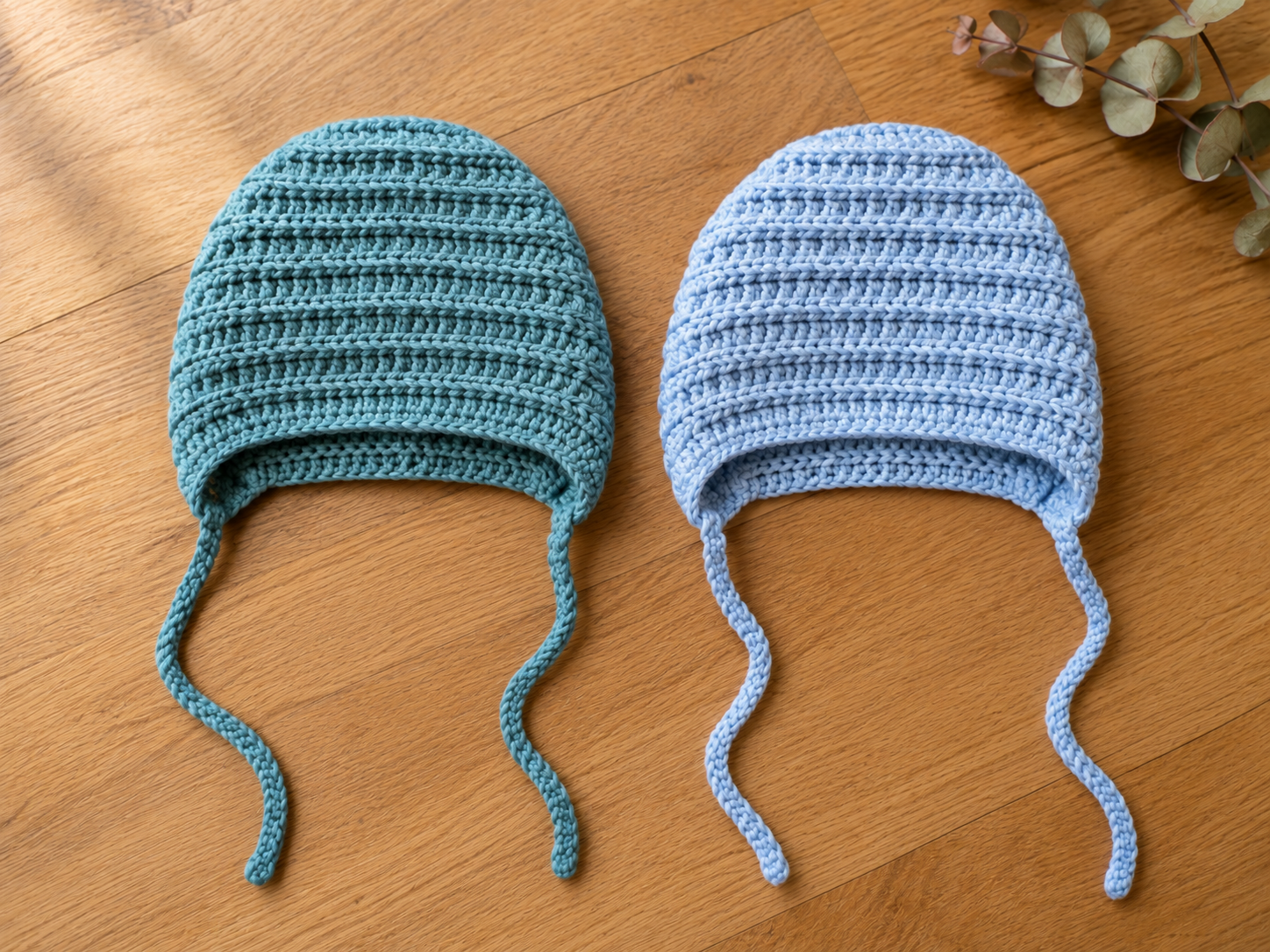 Two finished newborn crochet bonnets side by side on a warm oak wood floor, one in dusty teal and one in soft powder blue, both showing a ribbed half double crochet texture and long chain ties at each front corner