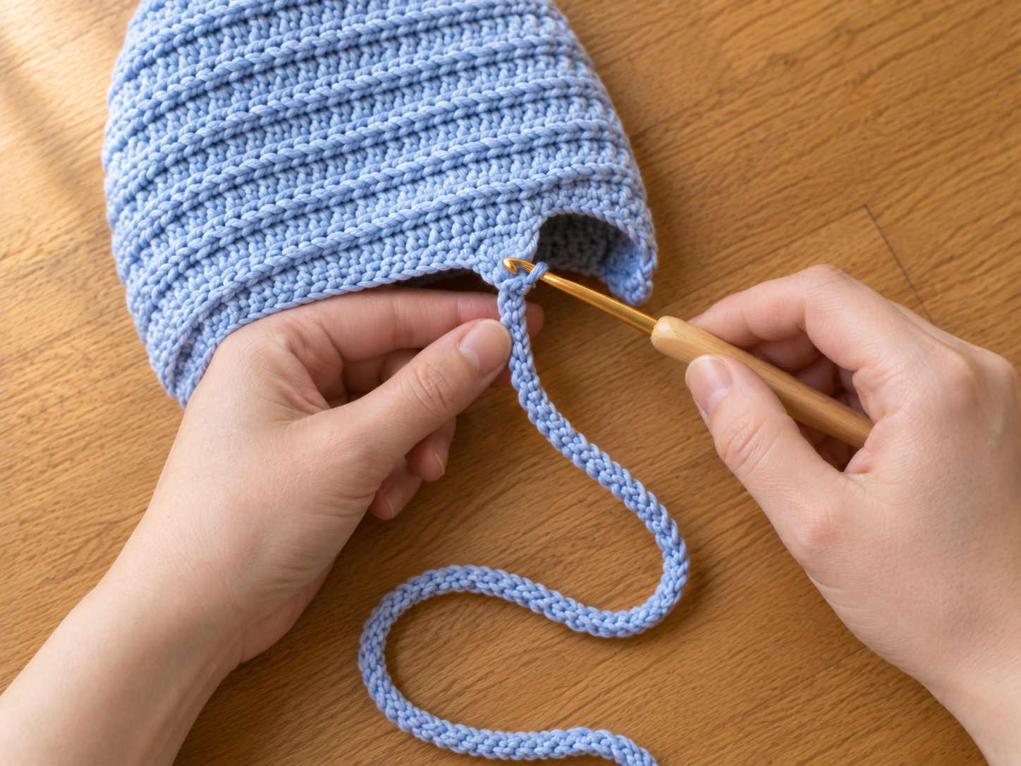 Close-up of hands working a long chain tie directly off the front corner of a powder blue crochet newborn bonnet with a 4 mm bamboo Clover hook, the ribbed hdc3lp texture visible on the bonnet body, on a warm oak wood surface