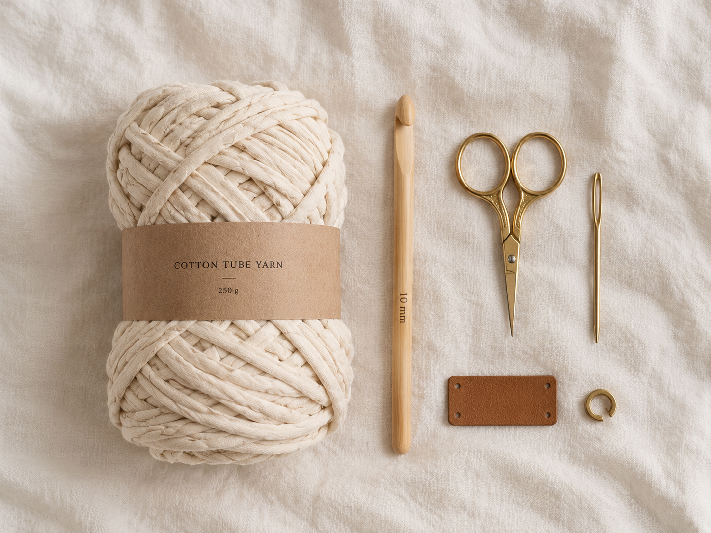 Overhead flat-lay of one natural cream cotton tube yarn skein, a 10mm bamboo crochet hook, small gold scissors, and a tapestry needle on a warm cream linen backdrop