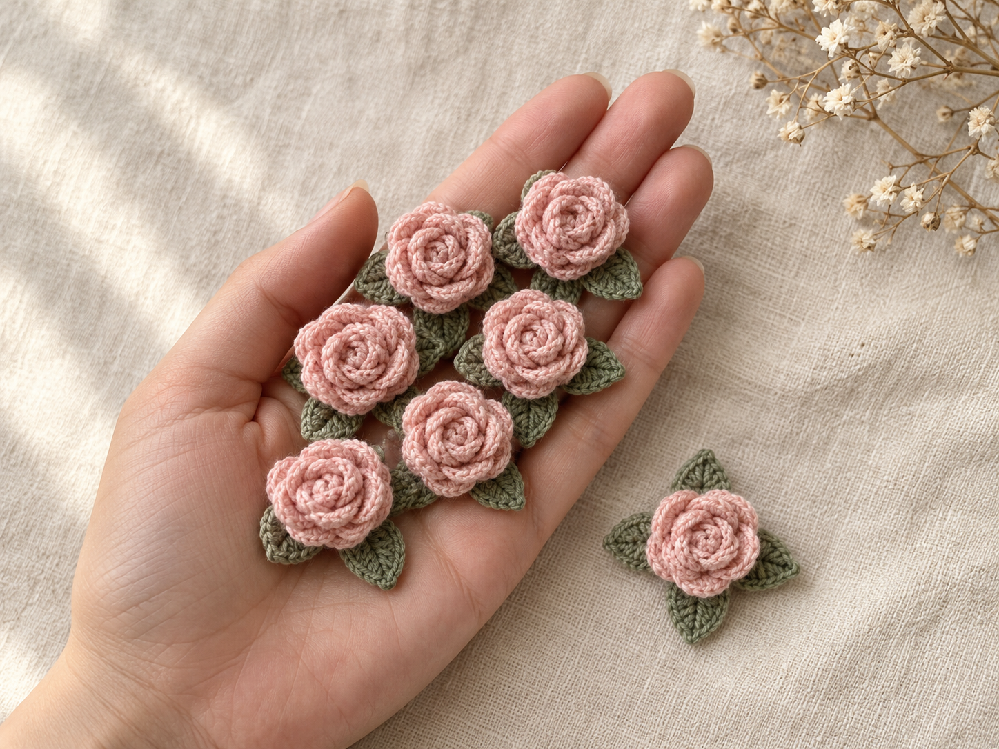 Seven blush pink crochet mini roses with sage green three-leaf bases resting on an open palm on cream linen