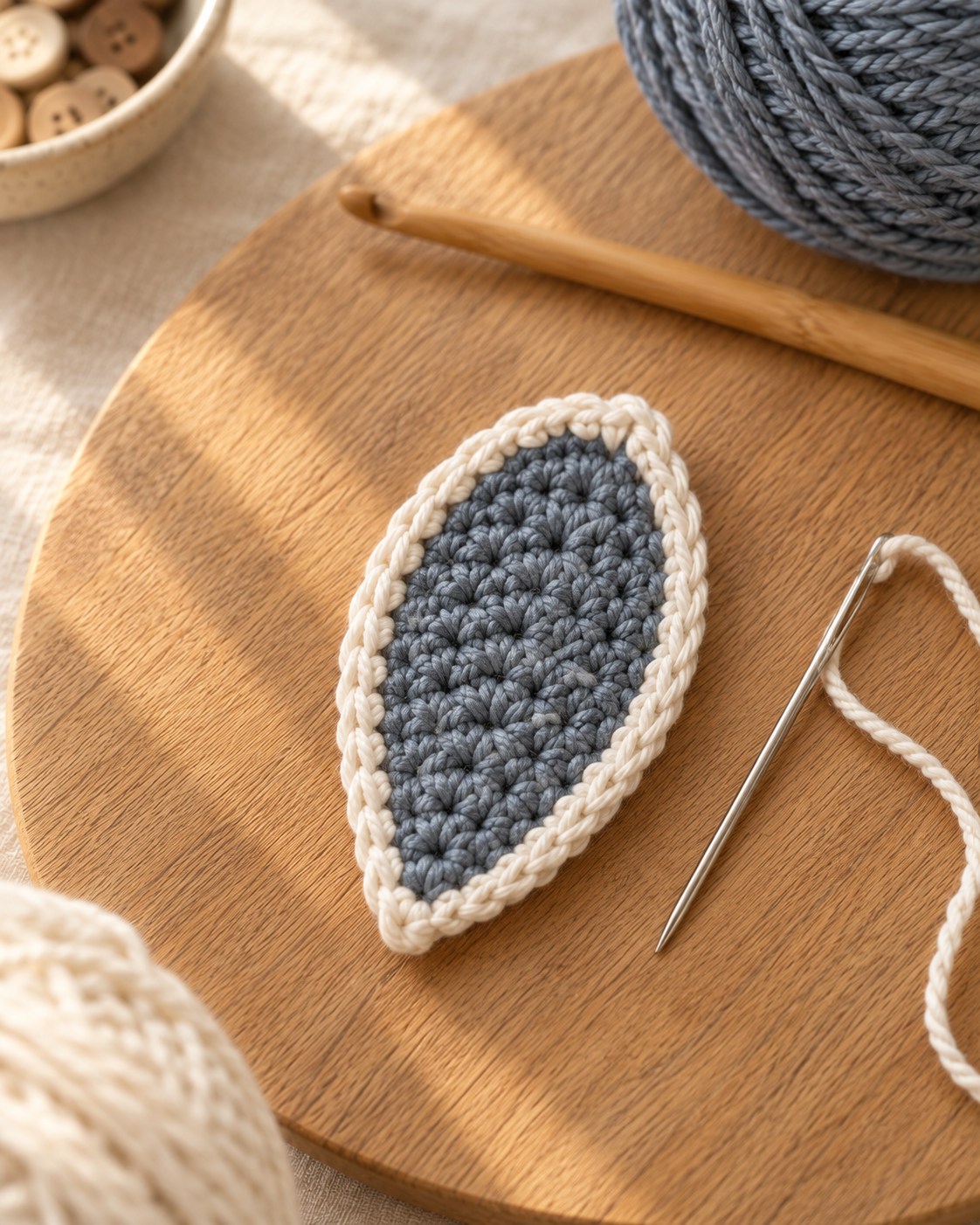 Ten slate blue-grey petals with cream trim arranged radiating around a cream center circle on a cream linen surface, pinned in place before sewing, a threaded tapestry needle resting beside