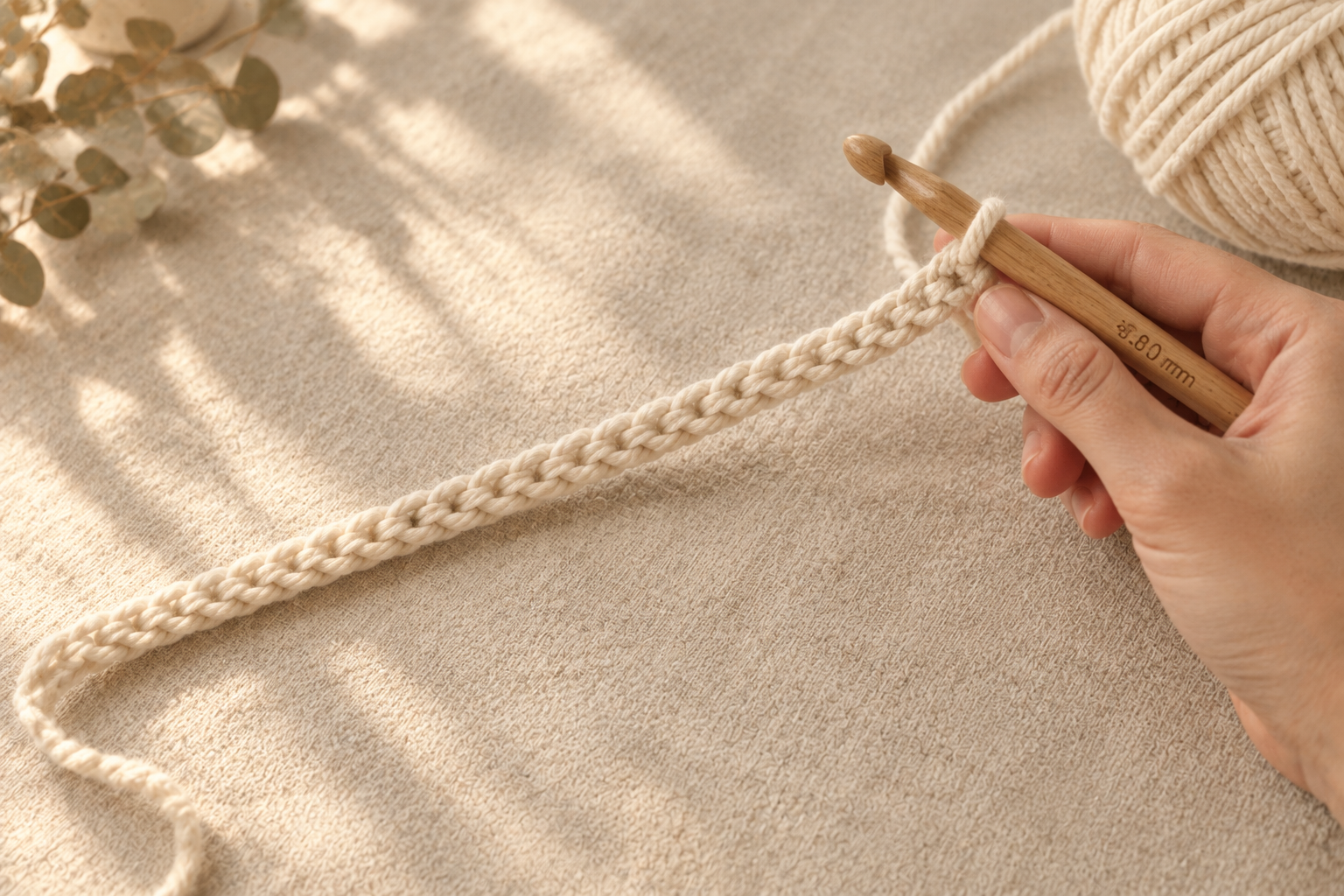 Hand holding an 8 mm oak crochet hook working a long cream foundation chain across a linen surface