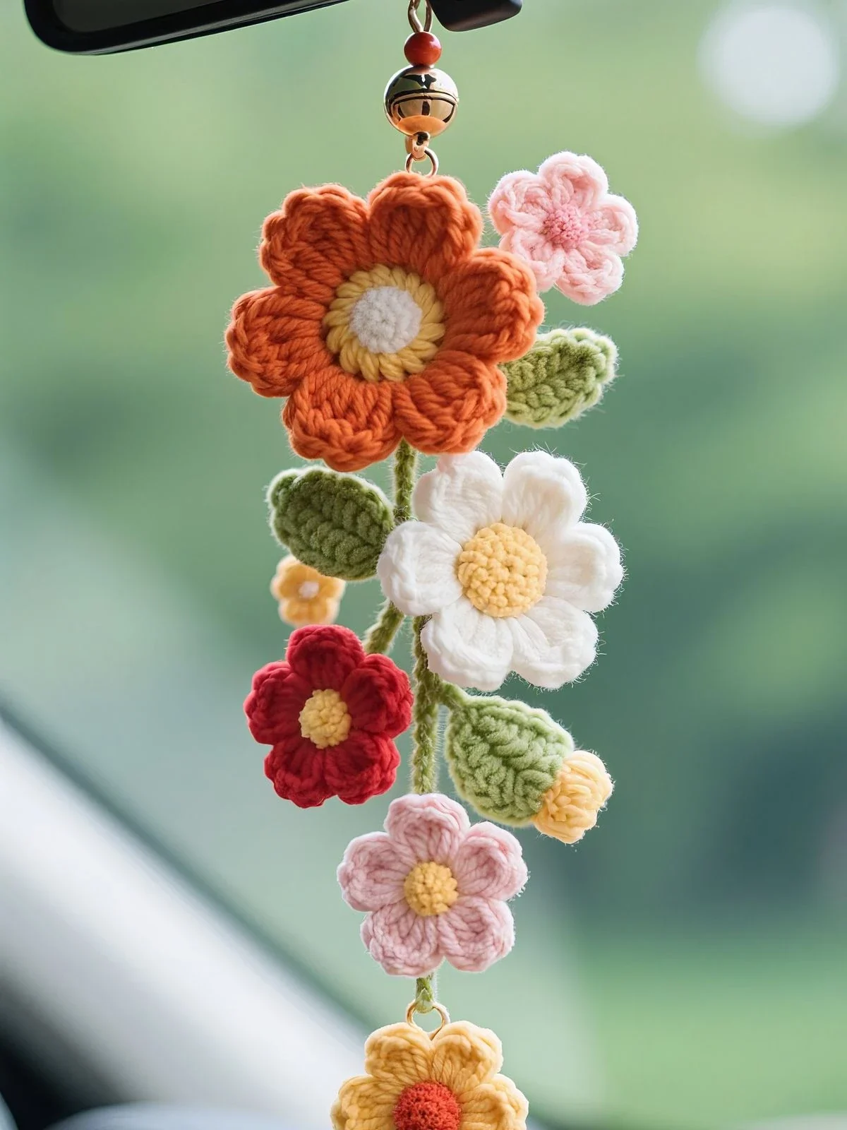 Crochet car mirror charm hanging from a rear-view mirror: a large orange flower with a yellow ring and white center at the top, then a pink flower, green leaves, a white daisy, a red flower, a small yellow flower, a yellow bud and a pink flower on a green stem, with a gold bell and red bead above and a yellow flower dangling at the bottom
