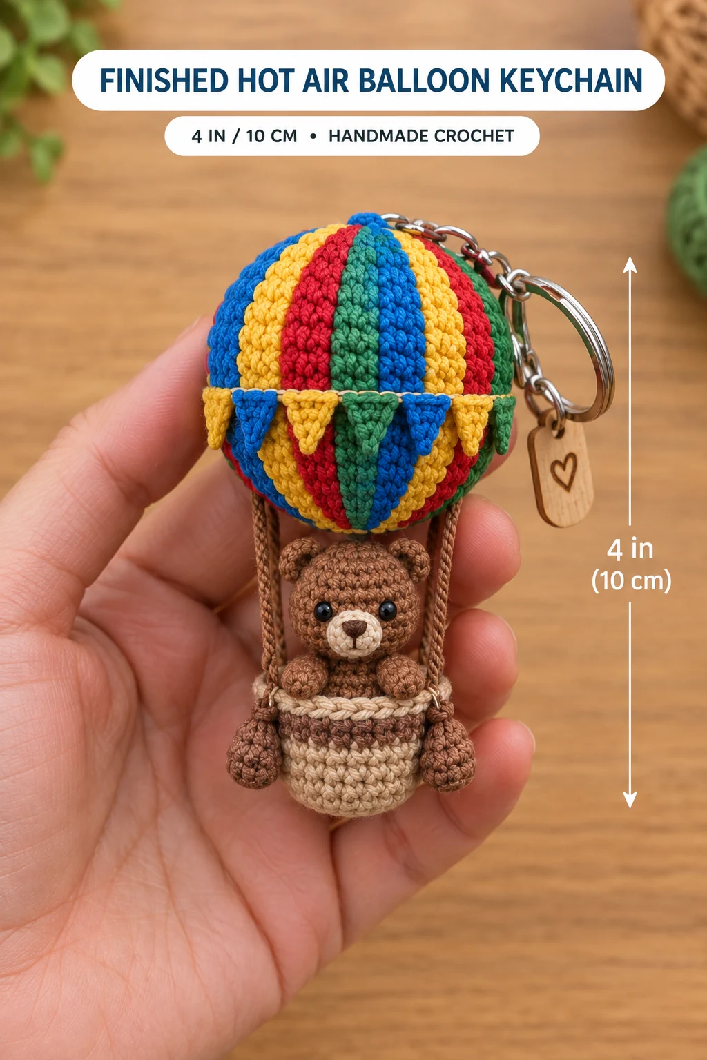 The finished crochet hot air balloon keychain held in a hand, about 4 inches (10 cm) tall, with a striped balloon, a band of bunting flags, a brown bear in a tan basket with sandbags, and a silver keyring with a small wooden heart tag