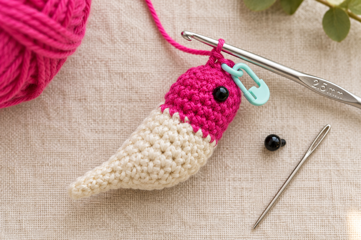 Overhead macro close-up of a mini crochet hummingbird body in progress — the magenta pink cotton head section finished with a small black safety eye already inserted and the cream cotton body section teardrop-shaped underneath, worked on a 2.5 mm silver crochet hook with a green locking stitch marker clipped into the working stitch, small ball of hot pink cotton DK yarn softly blurred in the upper-left corner, a second 6 mm black safety eye and a silver tapestry needle waiting beside on cream linen, warm natural window light