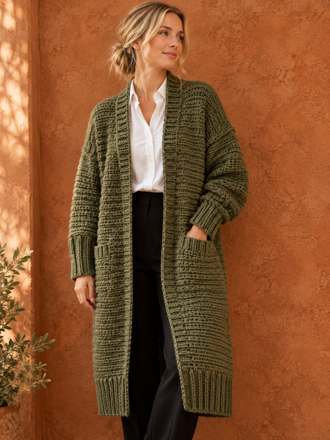 A blonde woman wearing a long oversized olive-green crochet duster cardigan over a white button-down shirt and black tailored trousers, showing the deep front pockets, ribbed cuffs, ribbed hem and the horizontal ribbed body texture, standing against a warm terracotta stucco wall with a small olive branch on the left, warm afternoon light, editorial cottagecore fashion photography