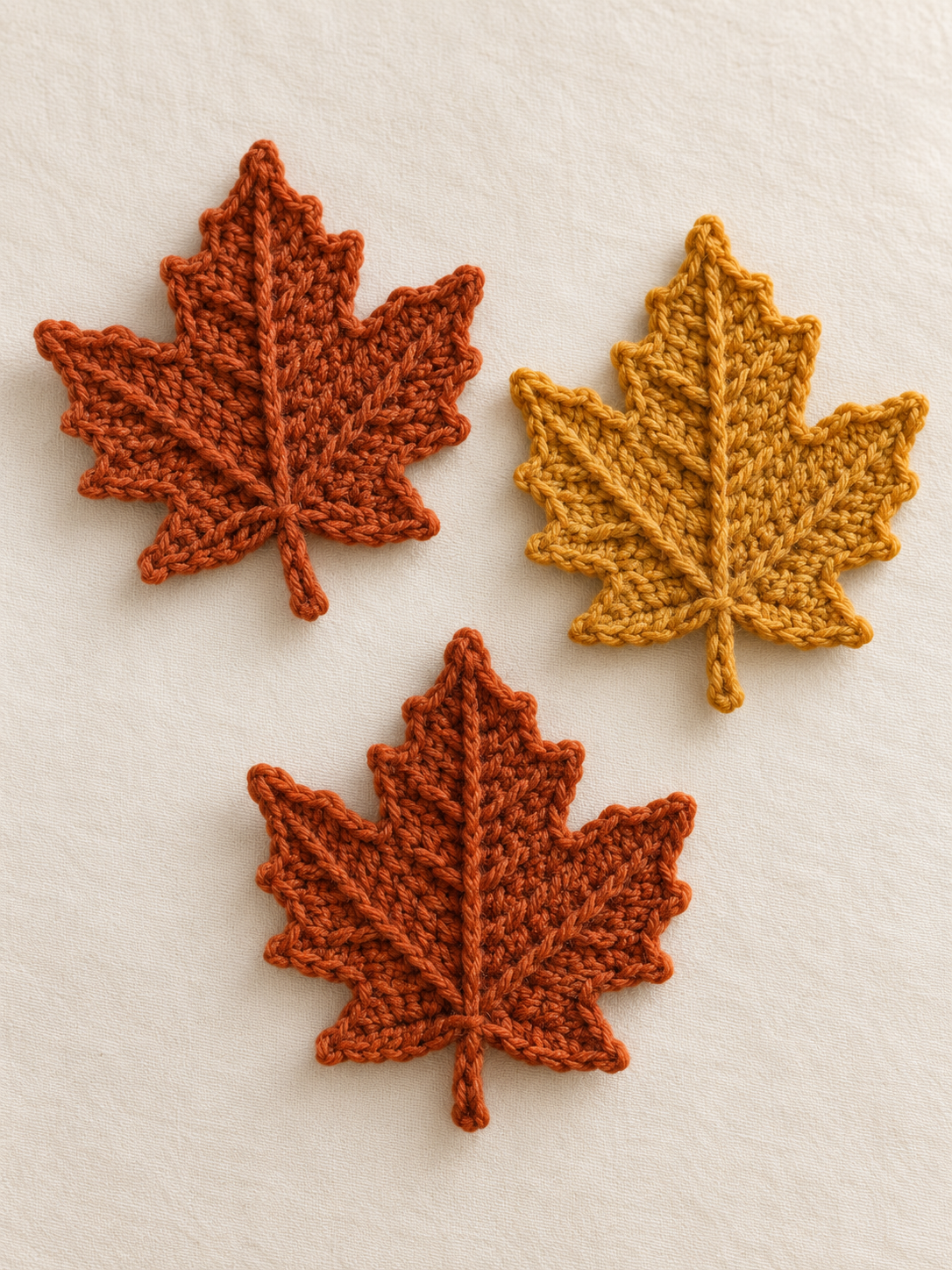 Three handmade crochet maple leaves arranged flat on a cream linen background — two rust-orange leaves in different sizes and one mustard-yellow leaf, each with the classic 5-point maple silhouette and a raised center vein worked in worsted-weight wool yarn, warm afternoon light, editorial cottagecore autumn craft-magazine styling