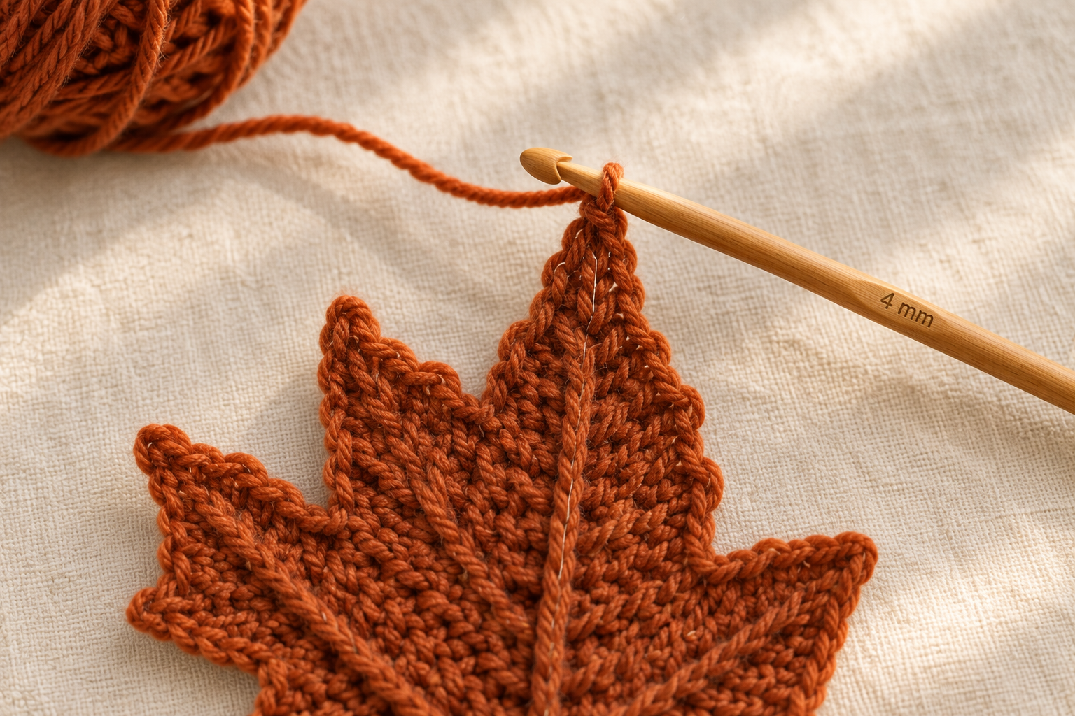 Overhead macro close-up of a rust-orange crochet maple leaf almost finished on a cream linen surface, the classic 5-point maple silhouette clearly visible with the raised center vein running down the middle, a 4 mm honey wooden crochet hook attached to the top of the leaf with the working yarn strand pulled out, a small ball of rust worsted-weight wool yarn softly blurred in the upper-left corner, warm afternoon window light, photorealistic craft-magazine tutorial detail shot, shallow depth of field