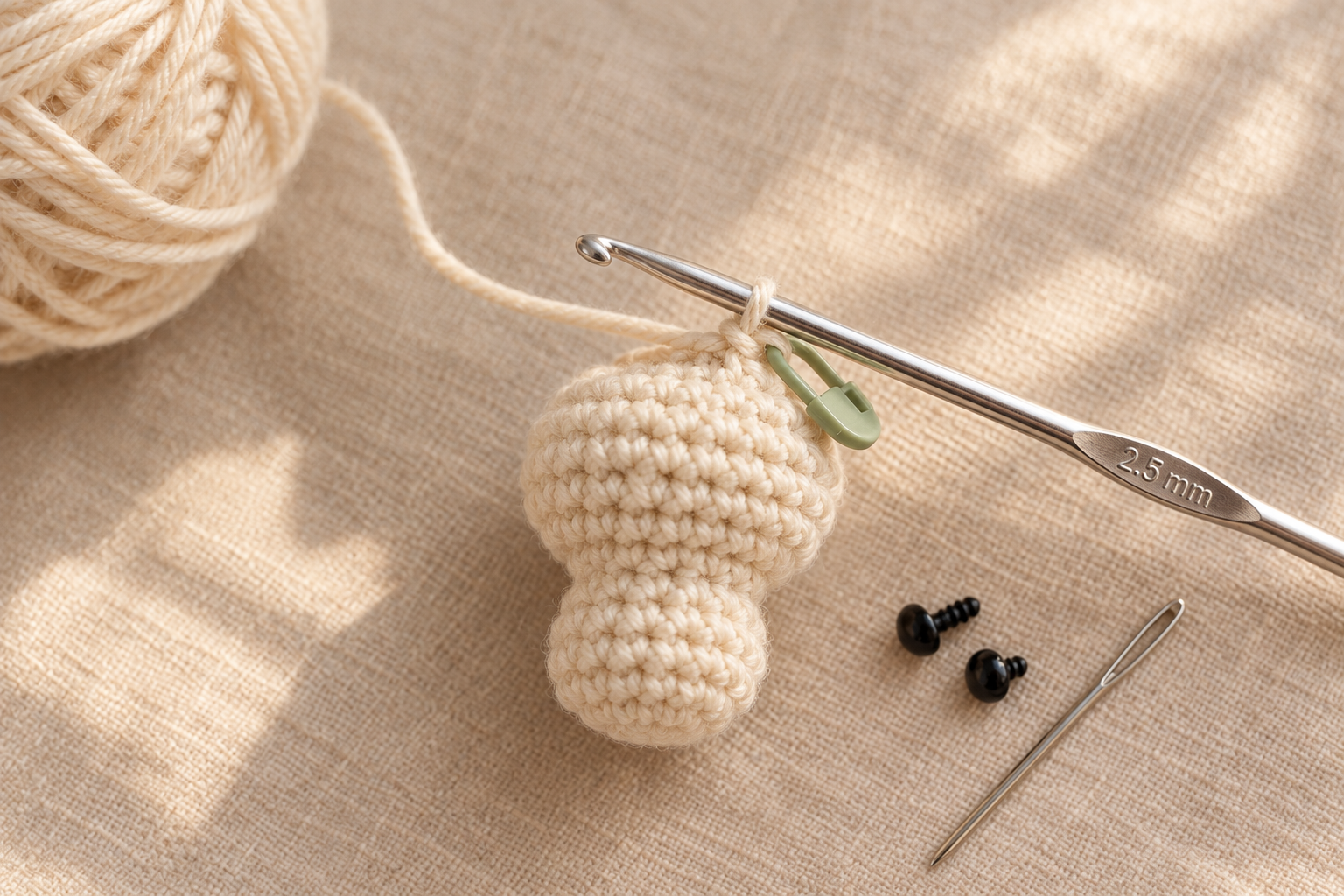 Overhead close-up of the cream cotton crochet mushroom body in progress on a 2.5 mm silver hook, small ball of cream cotton DK yarn softly blurred left, green locking stitch marker clipped into the working stitch, silver tapestry needle and two tiny 6 mm black safety eyes waiting beside on cream linen, warm natural window light, photorealistic craft photography, shallow depth of field