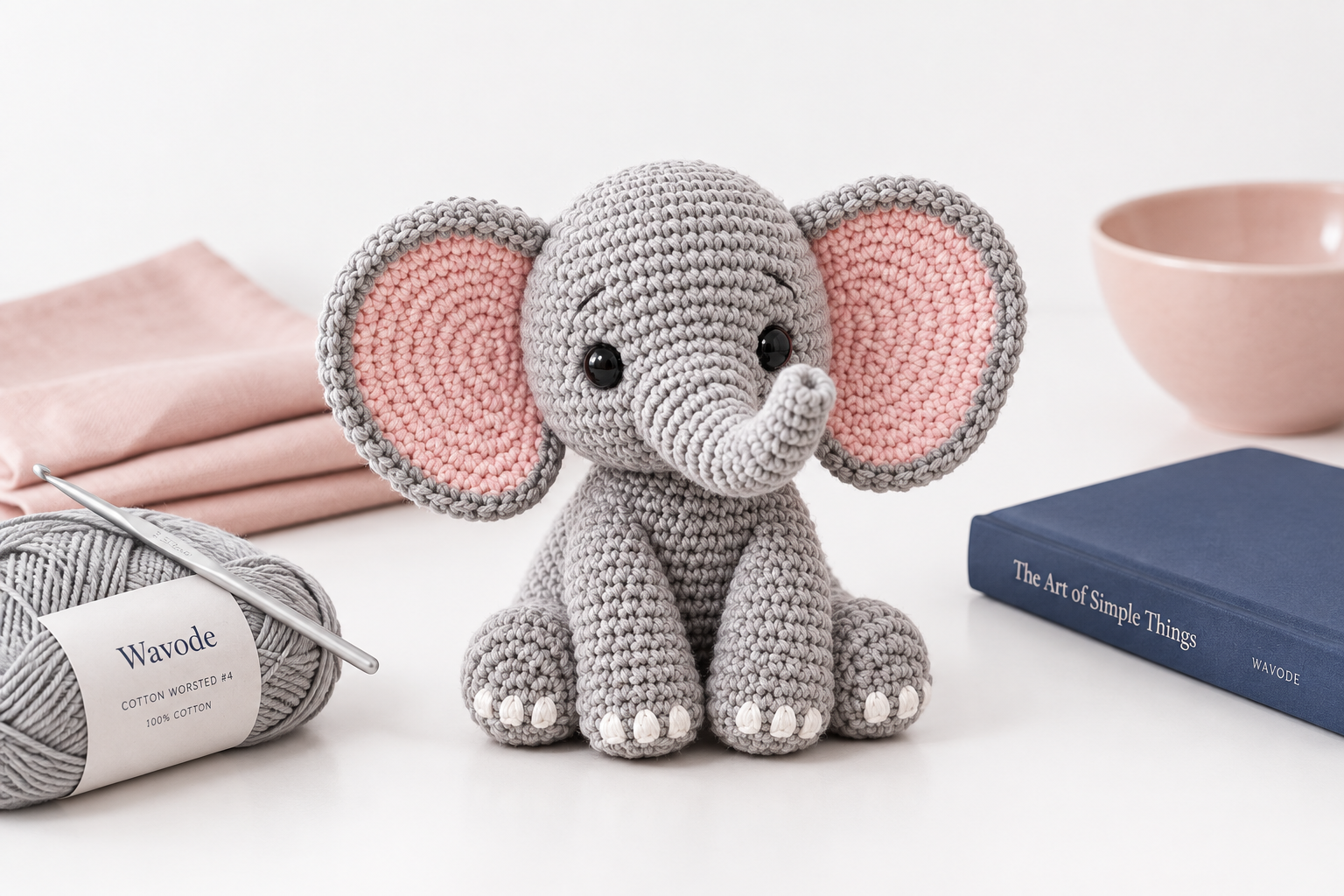 Grey crochet elephant amigurumi with big pink-lined ears, a curled-up trunk, black safety eyes and tiny white toenails, sitting on a pure white surface with a blush linen napkin, a navy hardcover book, and a Wavode grey worsted cotton skein beside it