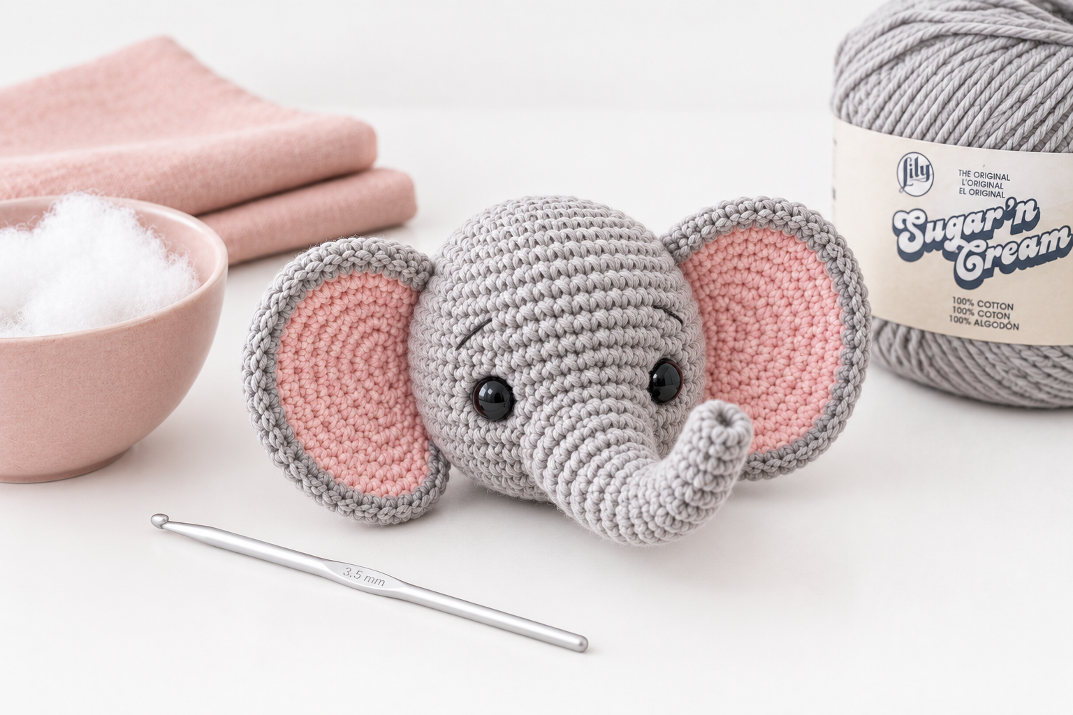 Finished grey crochet elephant head with pink-lined ears already attached and shiny black safety eyes, resting beside a blush ceramic bowl of polyfil stuffing, a Sugar n Cream cotton skein and a 3.5 mm aluminum hook on a pure white surface
