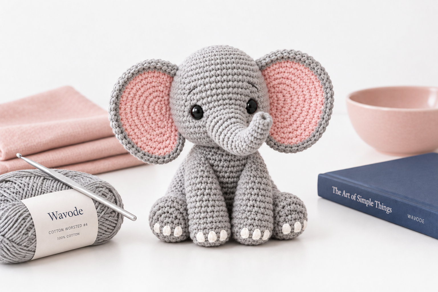 Grey Wavode cotton worsted #4 skein with a 3.5 mm aluminum hook, a finished grey elephant amigurumi with pink-lined ears, a blush linen stack, a blush ceramic bowl and a navy hardcover book on a pure white surface
