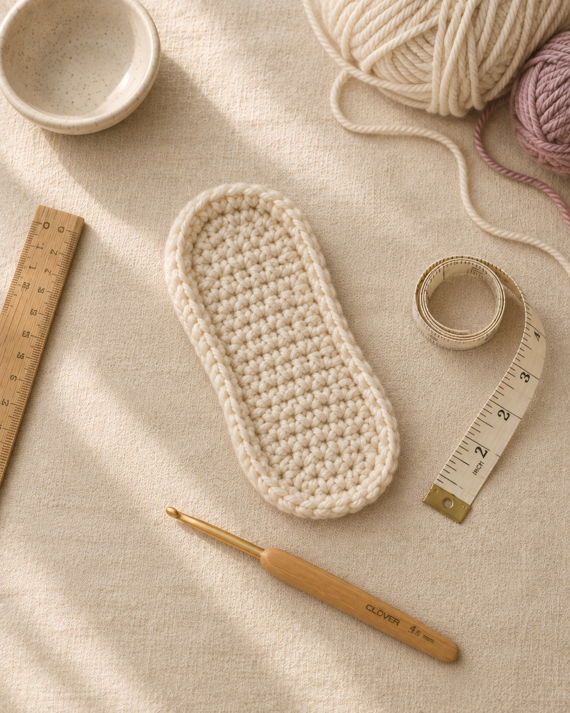 Flat-lay of one cream crochet oval bootie sole showing the classic elongated shape with a crisp BLO fold ridge, wooden ruler and 4 mm bamboo Clover hook and tape measure nearby on cream linen