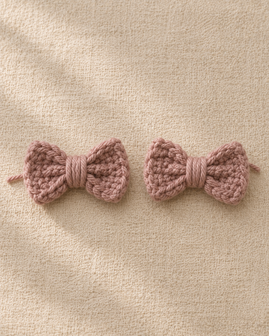 Close-up macro of two finished dusty rose crochet bows on cream linen, each showing the gathered rectangle pinched in the middle and the small mauve wrap around the center