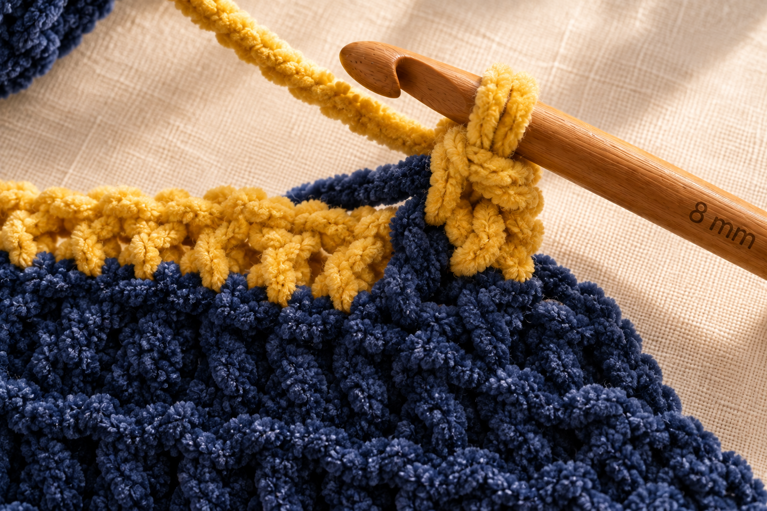 Extreme macro close-up of a chunky crochet color-change moment in progress at the end of a row, the last stitch of the navy blue band finishing with the yarn-over pulled through in bright mustard yellow Bernat Blanket chenille, both yarn strands visible with the navy dropping down and the mustard picking up on an 8 mm honey wooden crochet hook, cream linen background, warm afternoon window light, photorealistic craft-magazine tutorial shot