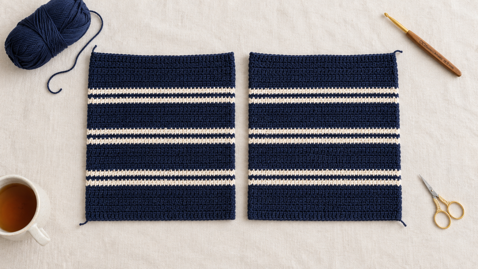 Overhead photograph of the front and back navy and cream striped crochet panels laid flat side by side on a cream linen surface with a navy yarn ball, wooden crochet hook, gold embroidery scissors and a mug of tea styled around them